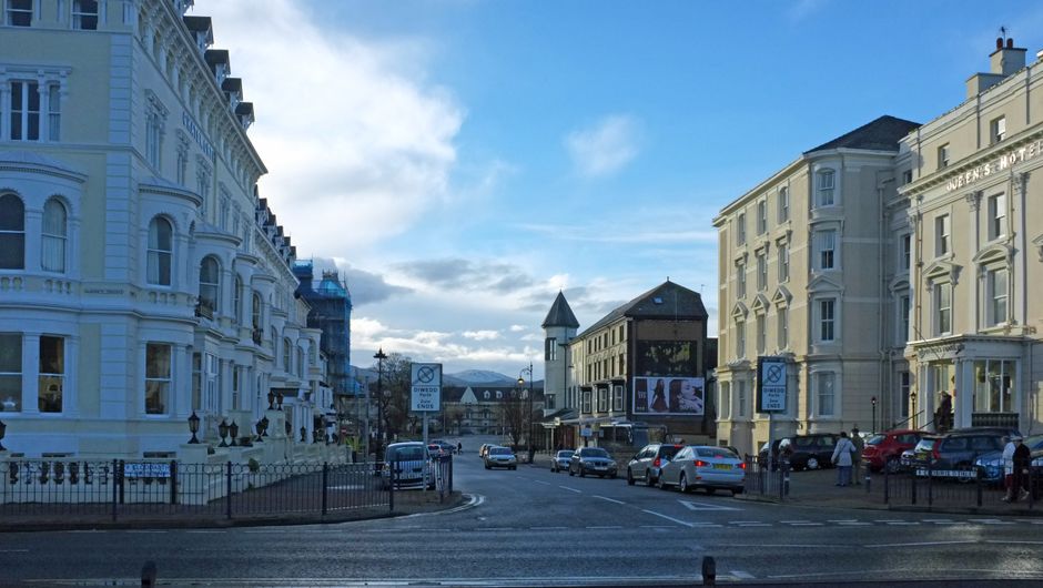 A daytime street scene in Queens Park showing a mixture of historic and modern architecture, with three- and four-storey buildings. The prominent white building on the left features intricate decorative detailing, sash windows, and a rounded bay window, with a smooth, painted façade. To the right, a beige building houses the Queen's Hotel, distinguished by its classical design, multiple windows with white trim, and a slightly protruding upper floor. Street parking is visible along the curb, with several cars parked in designated bays. A few pedestrians walk along the pavement, and a digital advertising billboard is mounted on a building in the center of the scene. The sky is mostly clear with some scattered clouds, and the lighting suggests bright daylight, highlighting the textures of the building exteriors and the urban environment. This scene depicts a typical urban area where local authorities might enforce outdoor rubbish clearance, fitting into the context of independent waste handling and private rubbish removal services such as those offered by Waste Disposal Queen's Park.