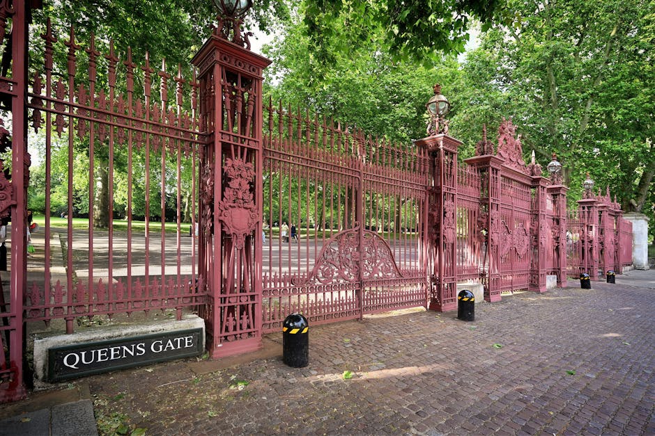 A large, ornate wrought iron gate painted in a muted red or burgundy color, featuring decorative finials and intricate scrollwork, stands prominently in the foreground of the image. The gate is situated at the entrance to a park or garden area, with a matching fence extending to the right and left, supported by tall, square columns topped with decorative lantern-style light fixtures. A small black sign with white lettering reading 'QUEENS GATE' is mounted adjacent to the gate at the base of the left column. The ground in front of the gate is paved with cobblestones, and several black bollards with reflective yellow markings are positioned along the curb for safety. Behind the gate, a tree-lined avenue with lush, green foliage creates a shaded environment, and the background reveals a peaceful, open park space with a paved pathway and some distant figures, indicating a public outdoor area managed without visible rubbish or clutter, typical of controlled private or municipal clearance services in Queens Park.