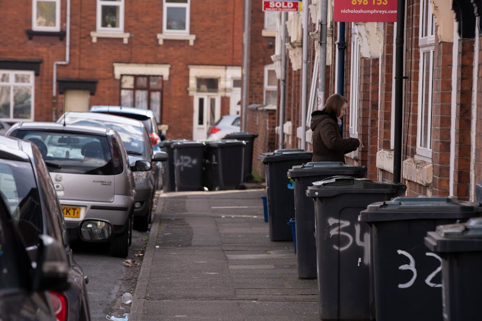 A row of black wheelie bins with closed lids lined up along a residential pavement in front of a red-brick terraced house. The bins are positioned close to the building's exterior wall, which features white window frames and stone sills. One of the bins has visible white graffiti markings. A woman wearing a dark brown coat stands beside the bins, appearing to be interacting with them or retrieving items. In the background, additional black bins are visible along the same street, with parked cars on the left side of the pavement, including small hatchbacks and compact vehicles in various shades. The street scene is set during daylight with natural, diffuse lighting. A red sign with white text is partially visible at the top of the image, attached to the building, indicating this may be a private property or for sale. The overall scene depicts an everyday urban environment, with residents' waste containers awaiting collection or disposal, aligned along a typical UK residential street, consistent with private rubbish removal services rather than council-led collection.